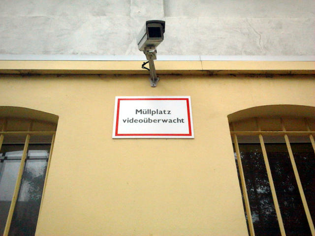 Überwachungskamera, an einer Wand montiert. Darunter ein Hinweisschild: 'Müllplatz videoüberwacht'.