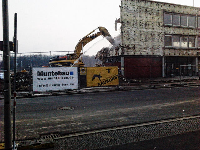 Foto eines Gebäudes, das gerade abgerissen wird. Davor ein Transparent am Bauzaun mit der Werbung 'Baukunst'.