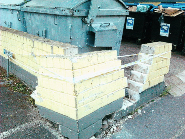 Ummauerter Platz für einige Müllcontainer neben einigen Parkplätzen. Eine Ecke der Mauer wurde mit großem Impuls von einem Autofahrer 'abgerissen'.